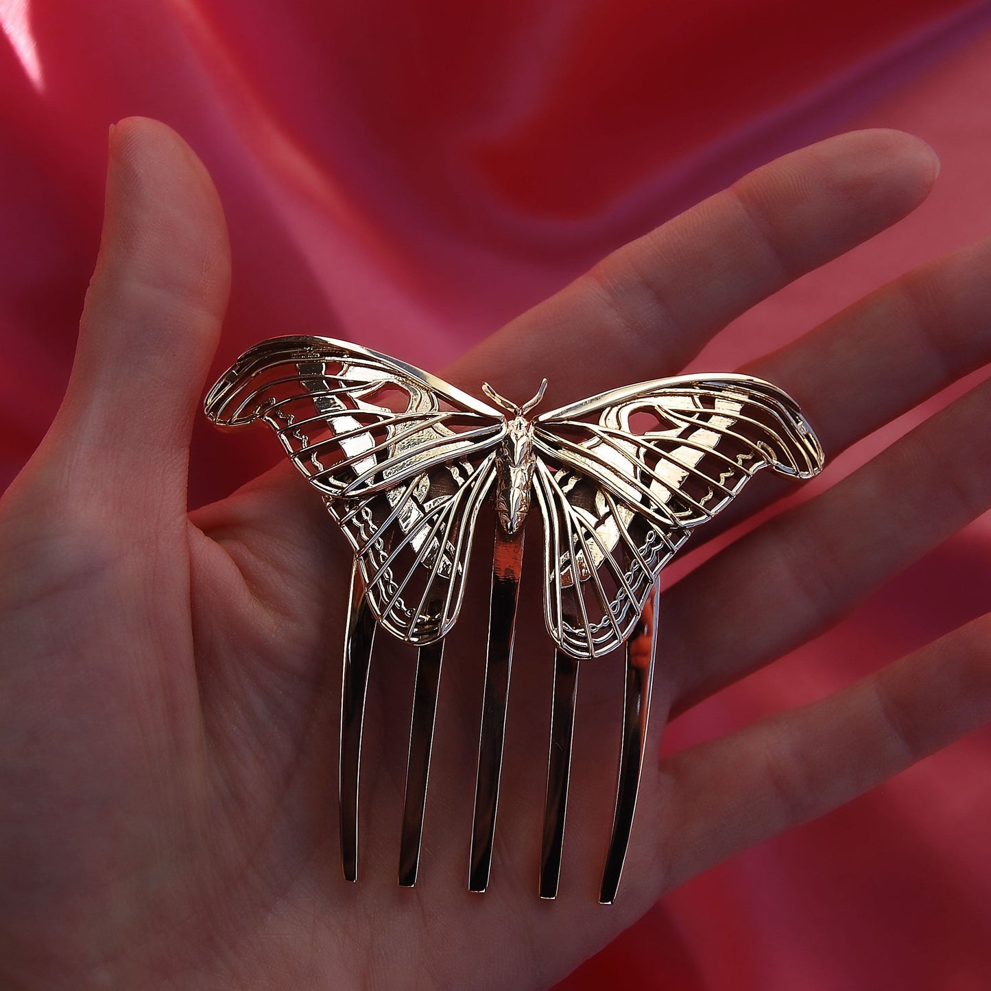 Hand holding a polished bronze Atlas Moth hair comb, showcasing handcrafted insect-inspired jewelry with detailed wing design and warm metallic finish.