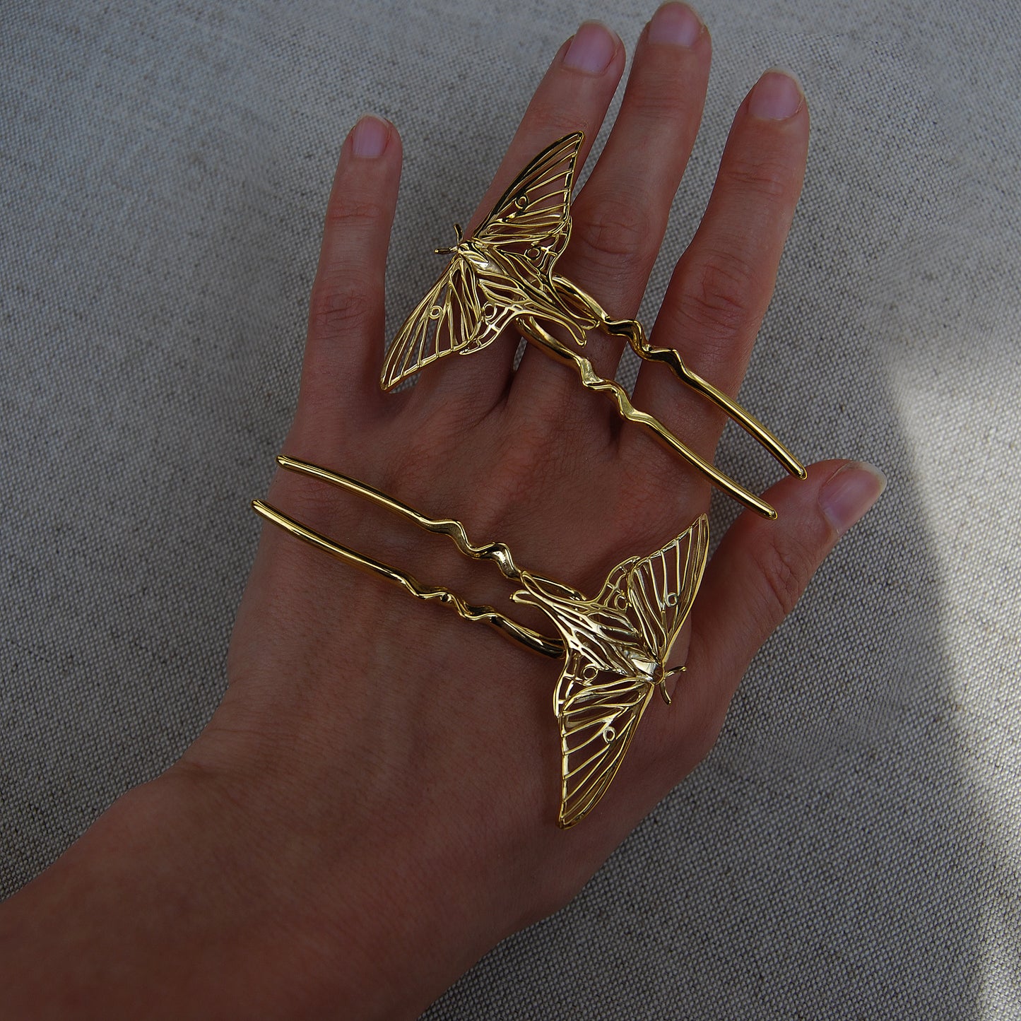 Hand holding two 14k gold-plated Luna Moth hair pins, showcasing symmetrical insect wing designs and polished metallic finish with intricate detailing.