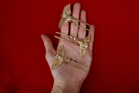 Hand holding 14k gold-plated Luna Moth hair pins, showcasing intricate insect wing design and polished metallic finish against a soft natural background.