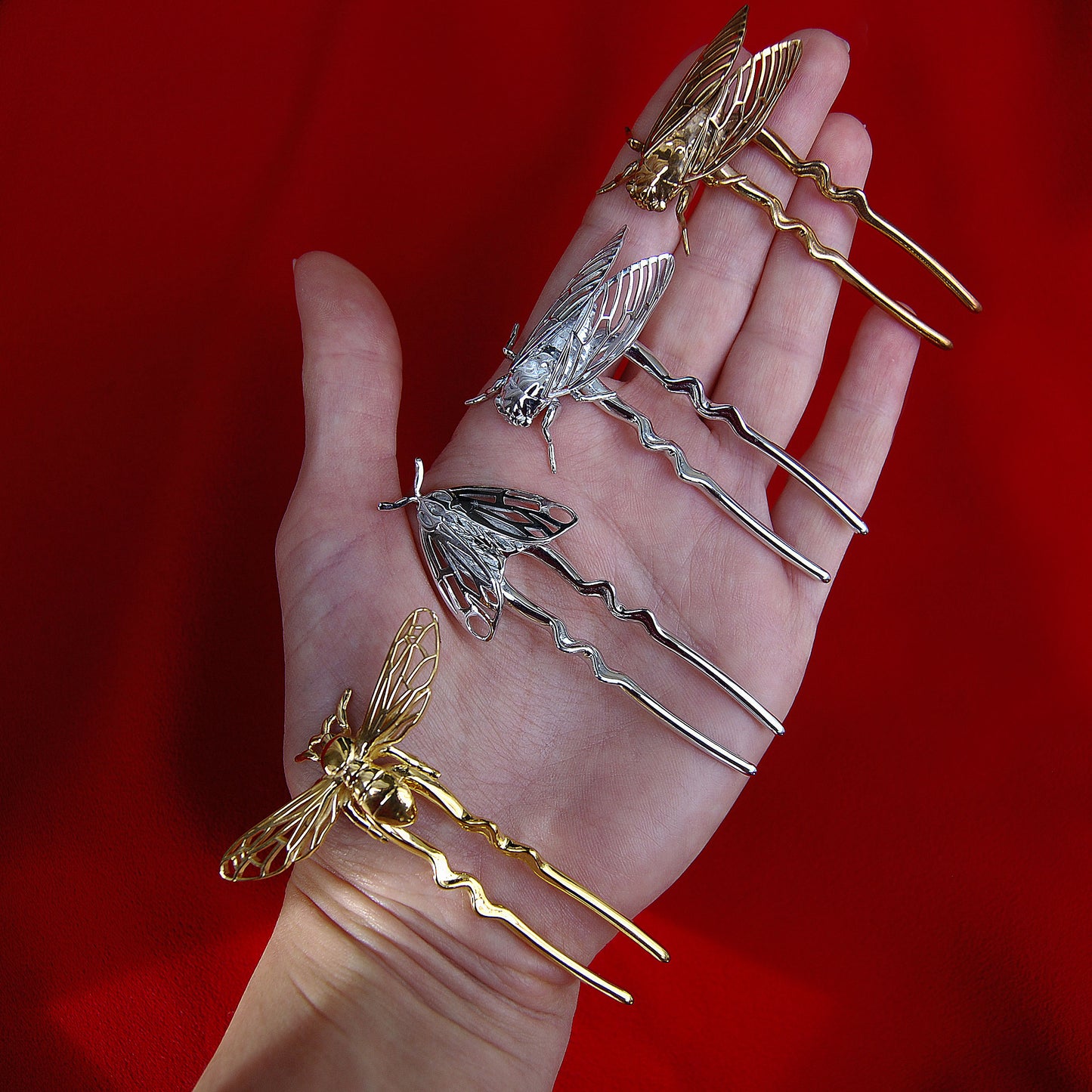 Hand holding a collection of insect-inspired hair pins, including a silver Tiger Moth, silver and bronze Cicadas, and a gold Bee, showcasing handcrafted sustainable jewelry in mixed metallic finishes.