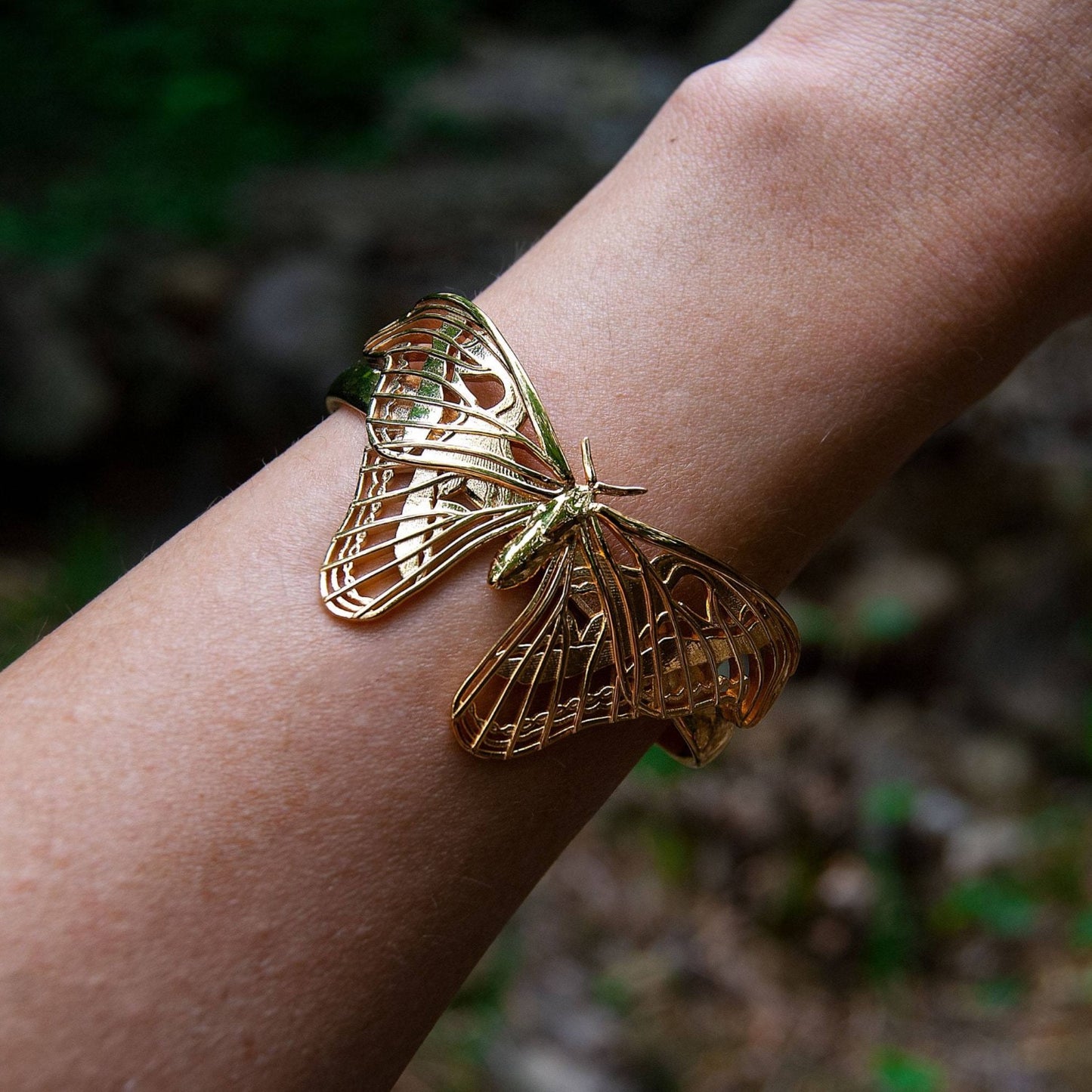 Atlas Moth Bracelet in 14k gold-plated brass displayed in a forest setting. The sculptural moth design gleams against earthy textures, with intricate wing details highlighted by natural light. A striking blend of organic inspiration and refined craftsmanship.