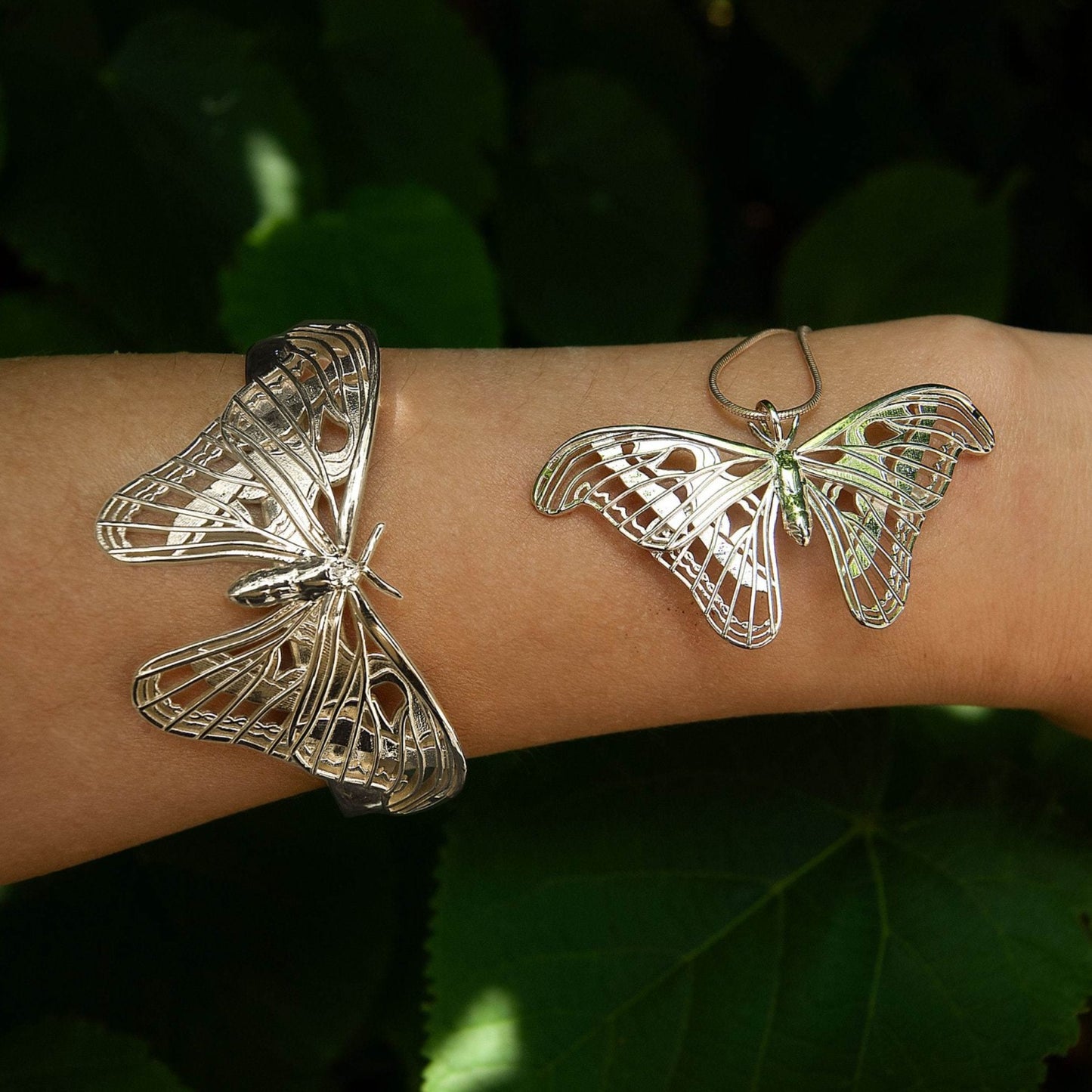 Sterling silver Atlas Moth pendant and matching bracelet worn on a hand, set against a lush nature background. The intricate moth design gleams in natural light, highlighting its delicate wing details and sculptural elegance. The organic setting enhances the jewelry’s connection to the natural world, emphasizing its inspiration from one of Earth’s largest and most beautiful moths
