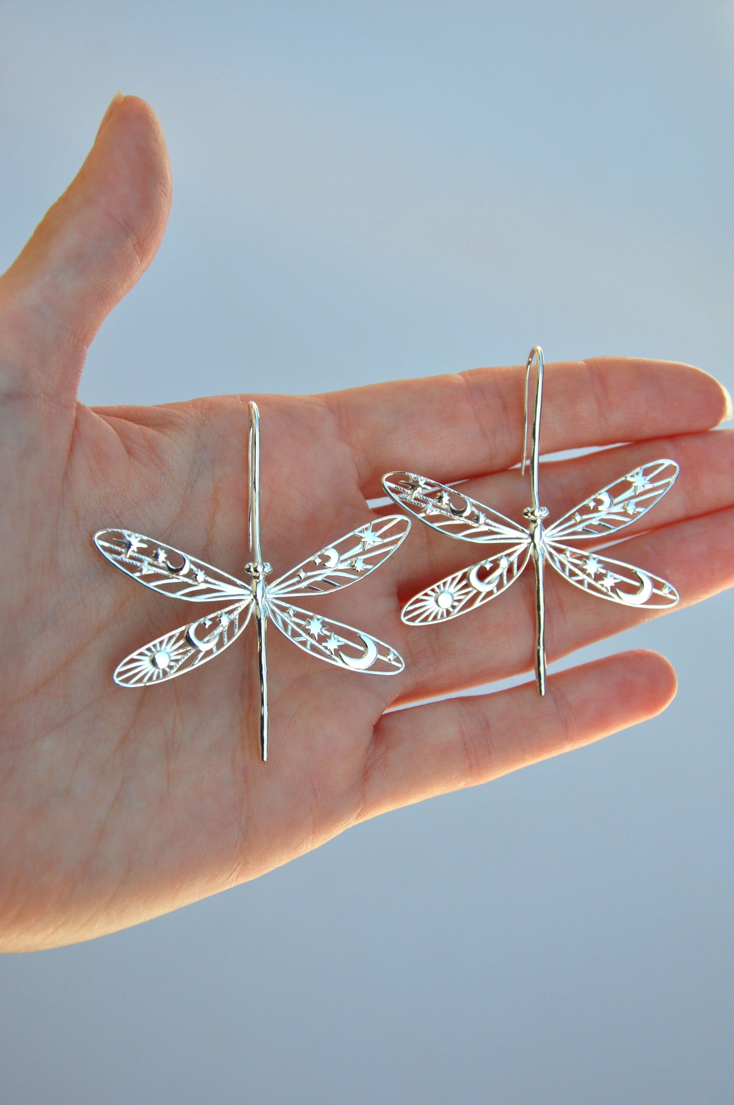 Hand holding two damselfly earrings with delicate celestial cut‑out wings, including stars, moons, and sun shapes