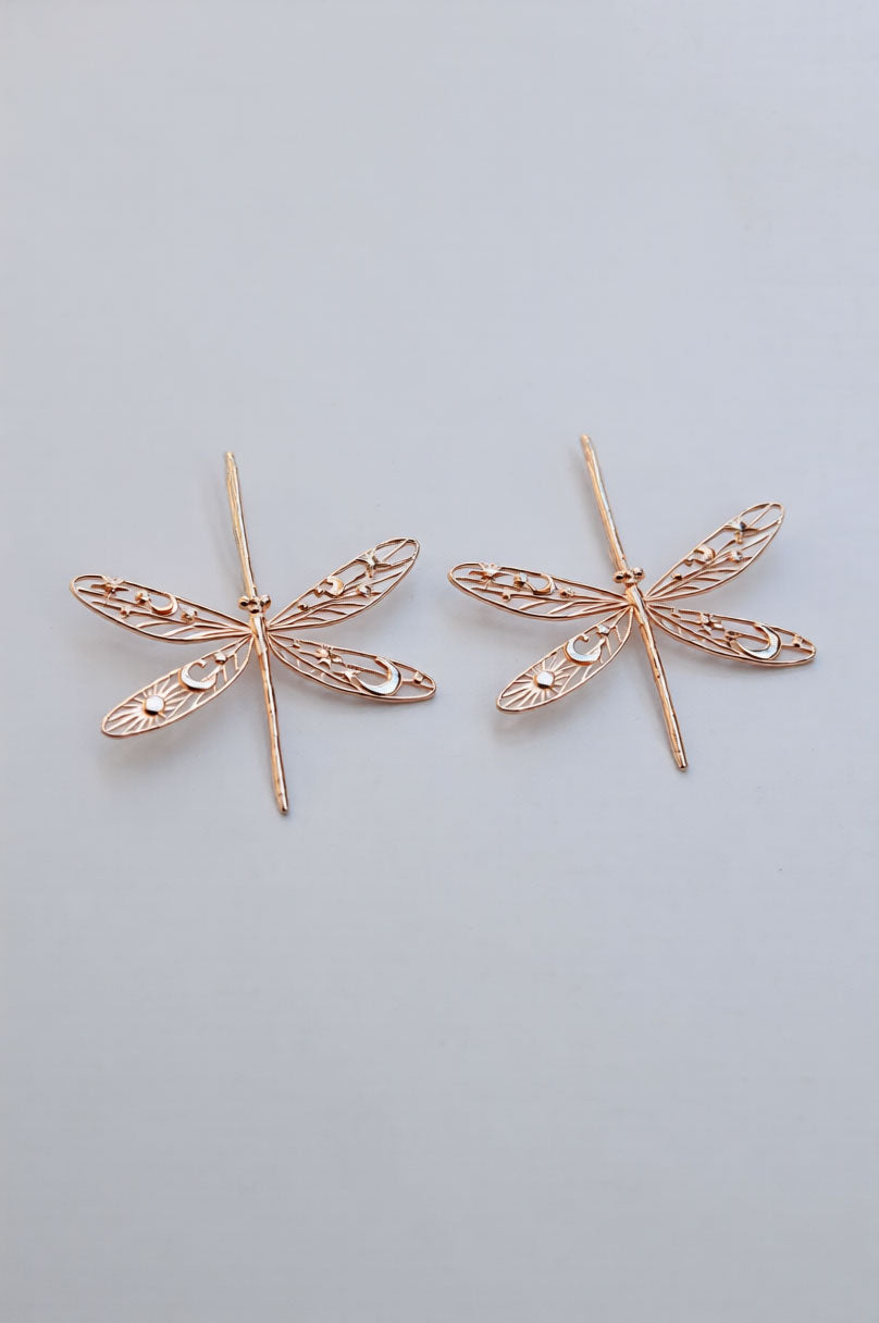 Rose‑gold damselfly earrings with filigree celestial wings decorated with star, moon, and sun cut‑outs.