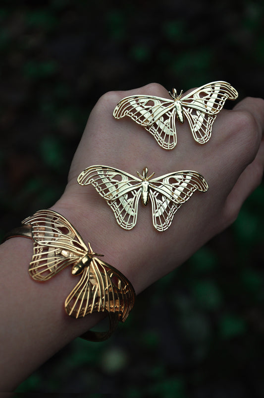 A close-up image of a gold-colored moth pendant being worn on a person's wrist, with blurred greenery in the background.