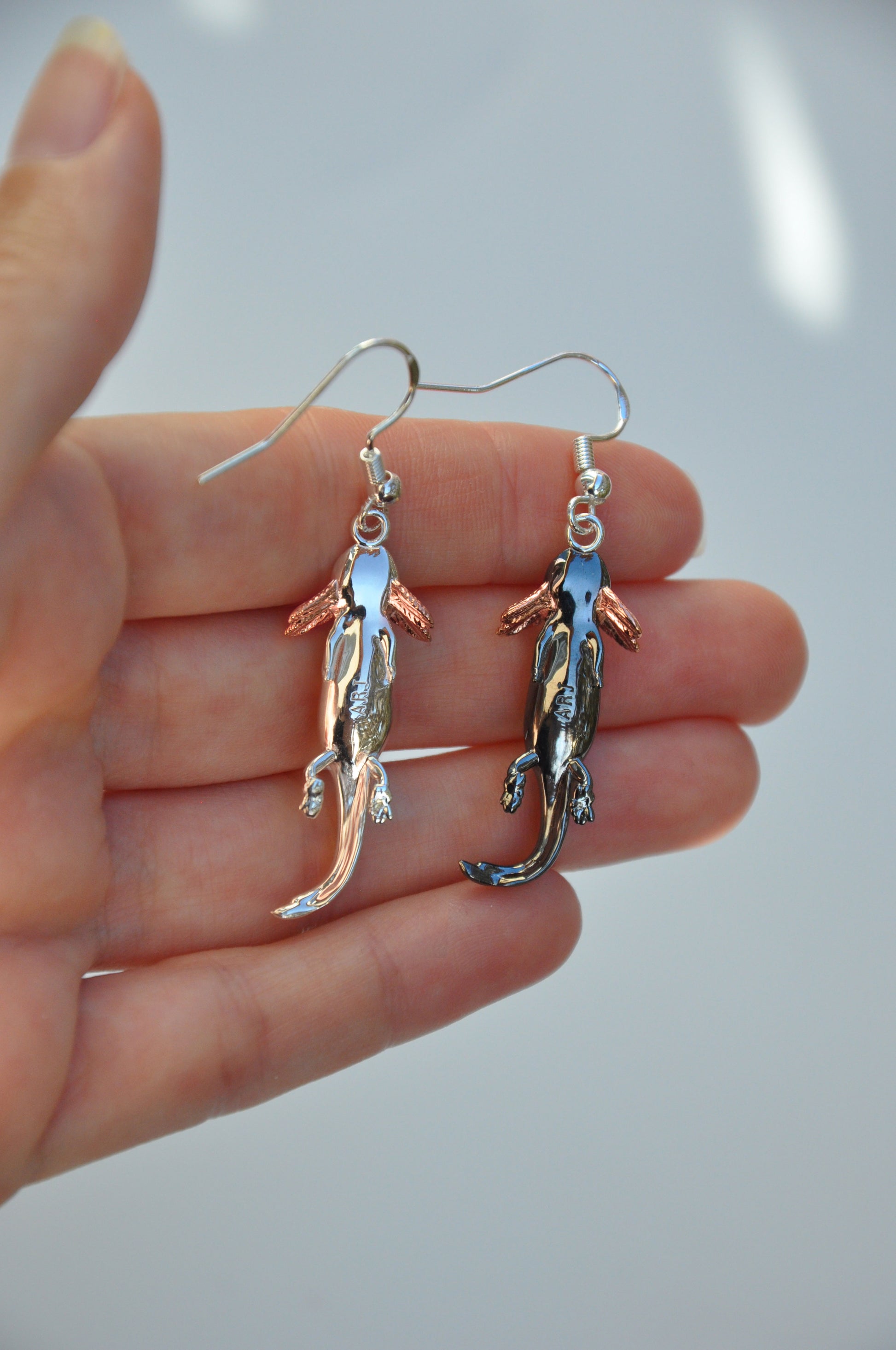 Close-up of the Axolotl Drop Earrings held in hand, front-facing to emphasize the expressive axolotl design and rose gold-plated gills.