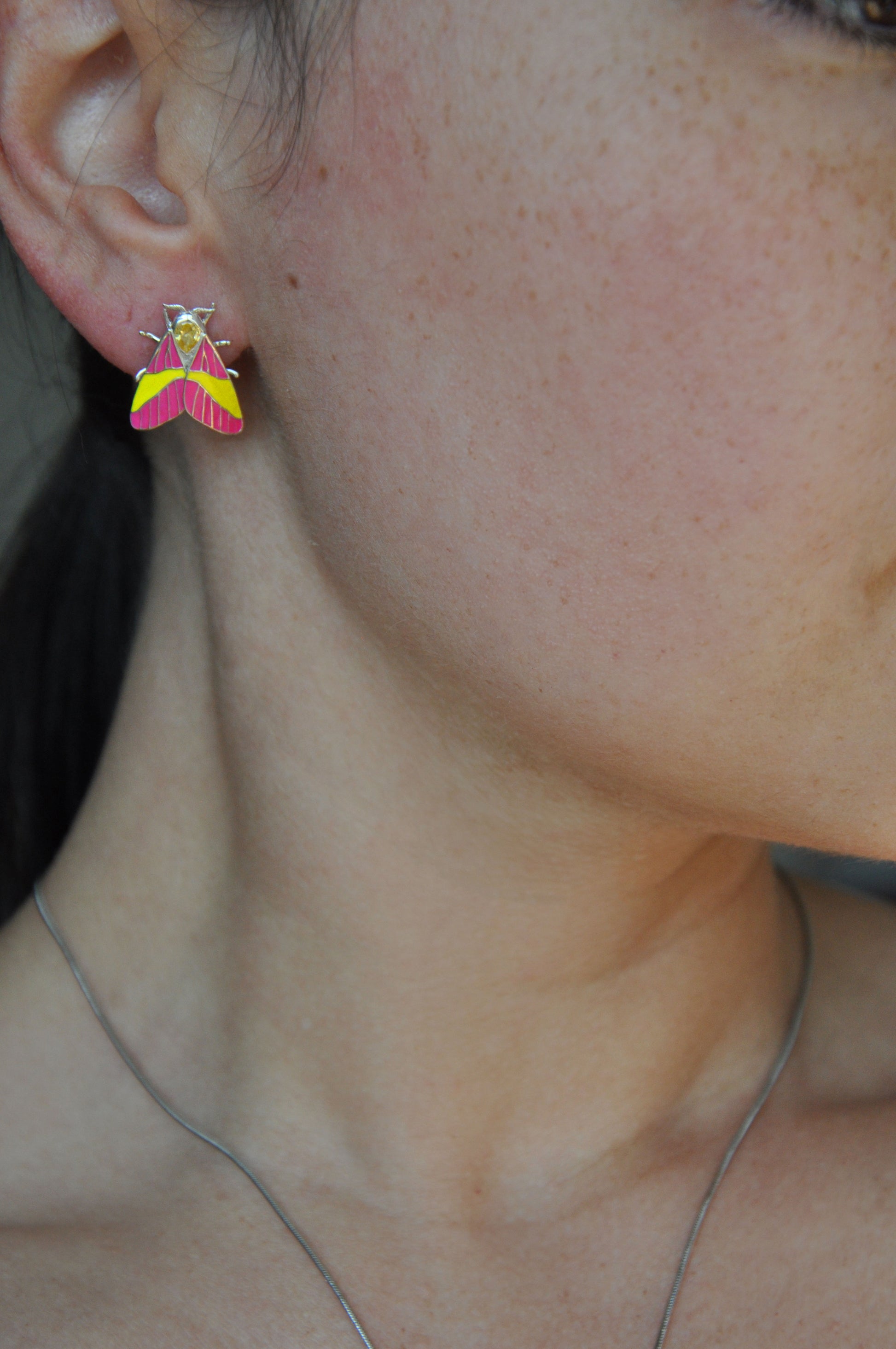 Model wearing Rosy Maple Moth earrings in sterling silver with pink and yellow enamel wings, blue gemstone eyes, and yellow pear-shaped crystals