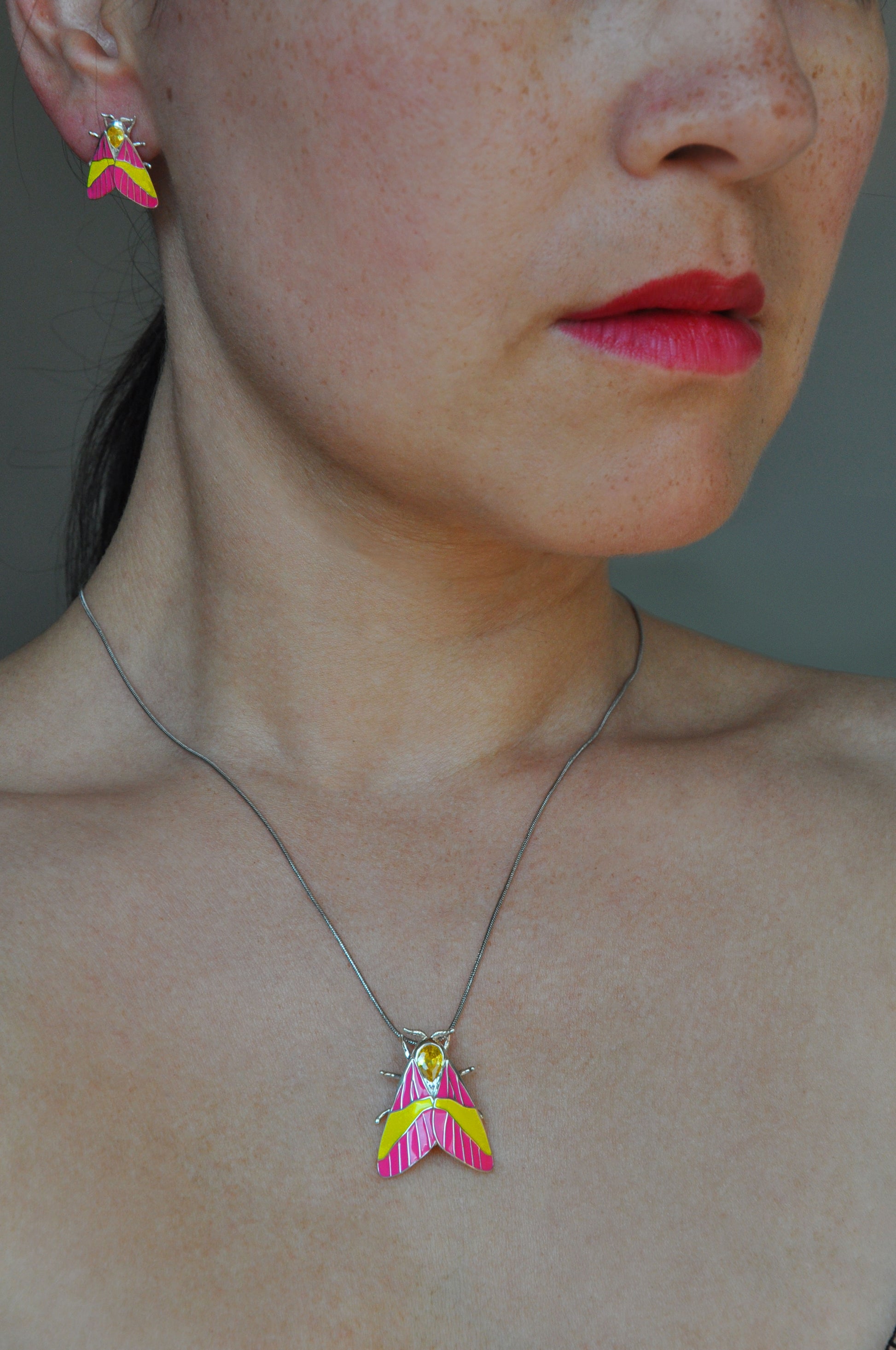 Model wearing Rosy Maple Moth pendant and earrings in sterling silver with candy-colored enamel, blue gemstone eyes, and yellow crystal accents