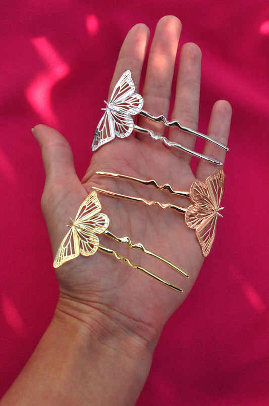 Butterfly-shaped hair pins in gold and silver held in a hand against a pink background