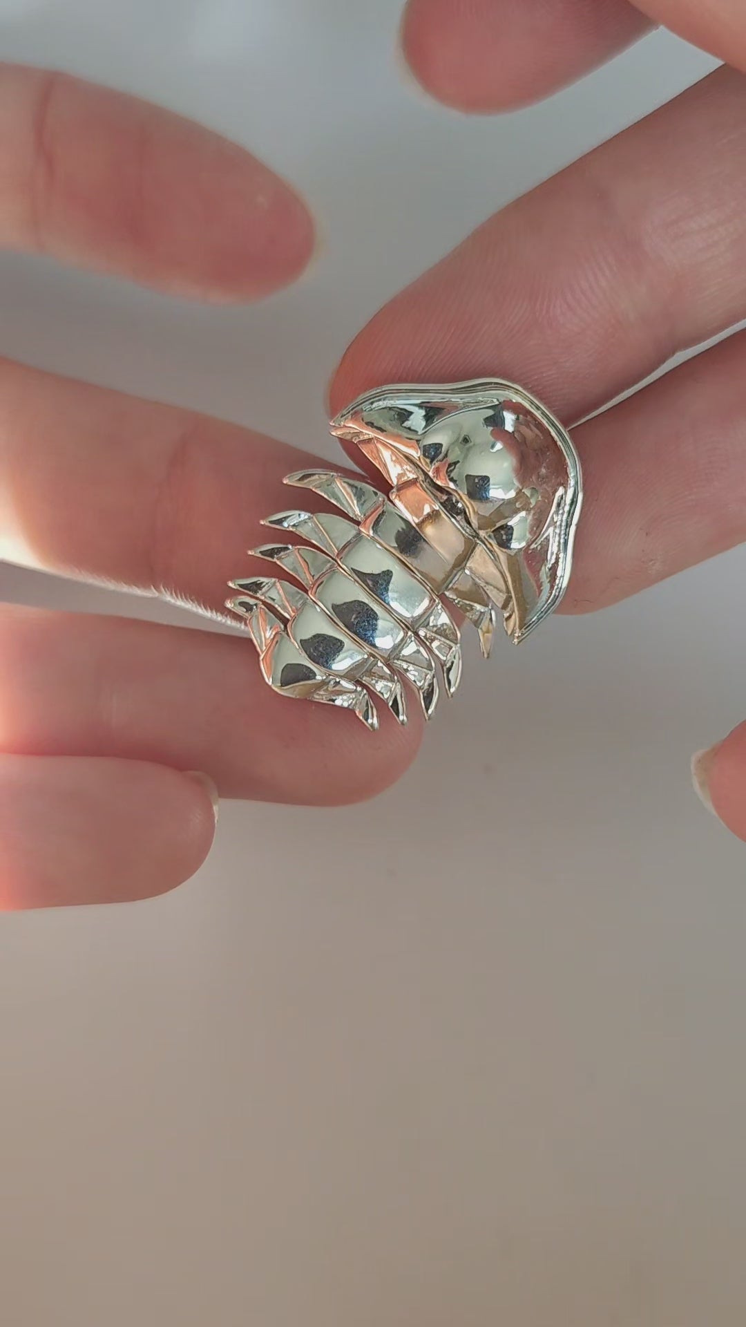 Hands gently manipulate the sterling silver articulated trilobite pendant against a white background, showcasing the fluid motion of its segmented body. Each joint flexes smoothly, highlighting the pendant’s lifelike mobility and intricate craftsmanship inspired by ancient marine fossils.
