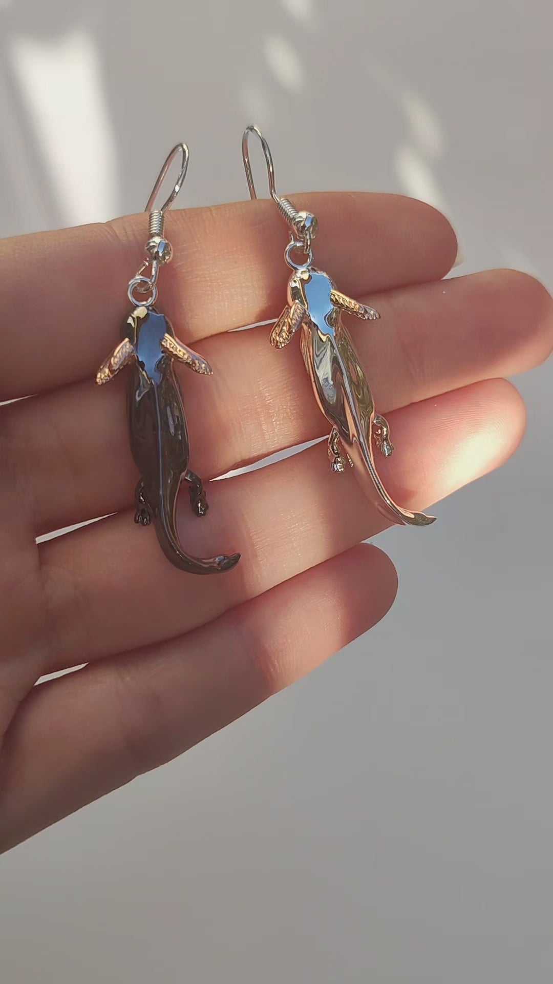 Video showing the Axolotl Drop Earrings held in hand, rotating to reveal their sculpted details, contrasting finishes, and shimmering rose gold gills.