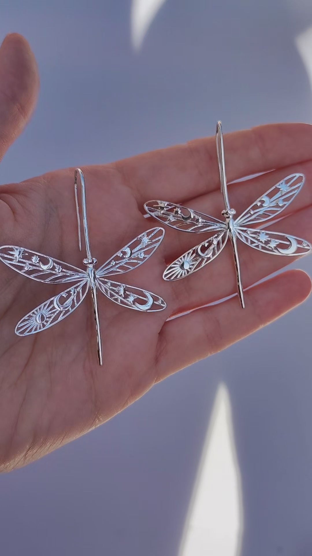 Video of a hand holding celestial damselfly earrings, showing the detailed cut‑out wings with star, moon, and sun motifs as they move in the light