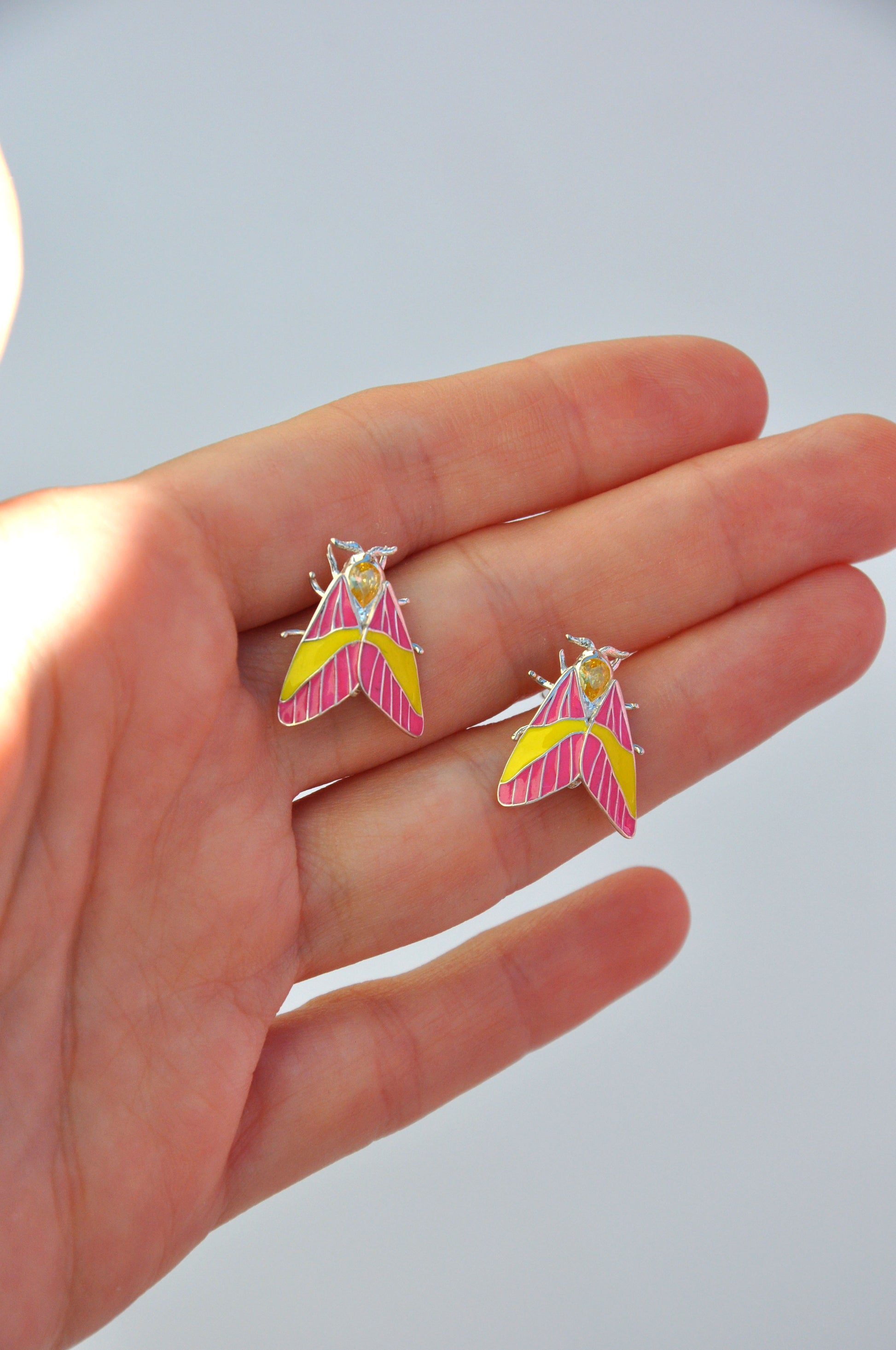 Rosy Maple Moth earrings held in hand, showing scale and enamel craftsmanship