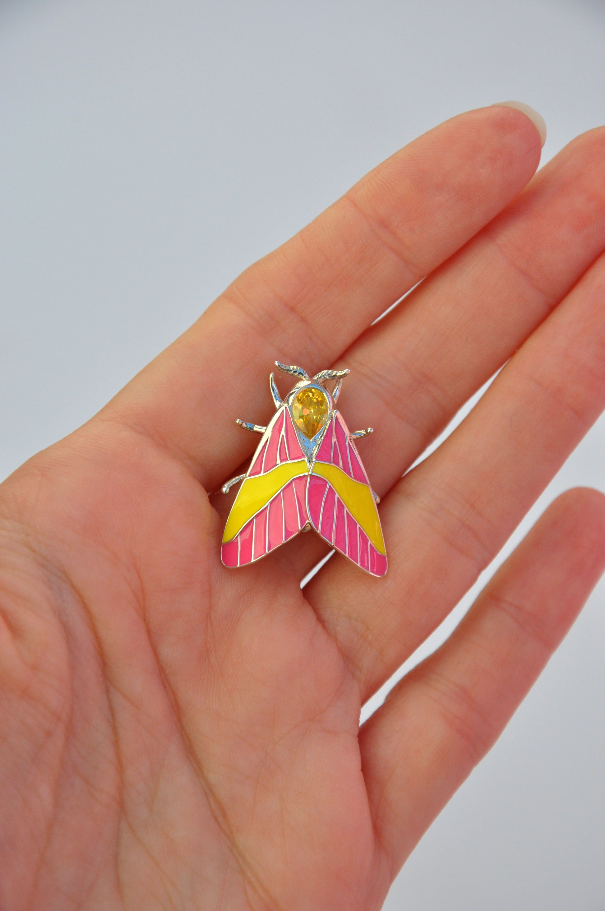 Rosy Maple Moth pendant held in hand, showing scale, enamel colors, and gemstone sparkle