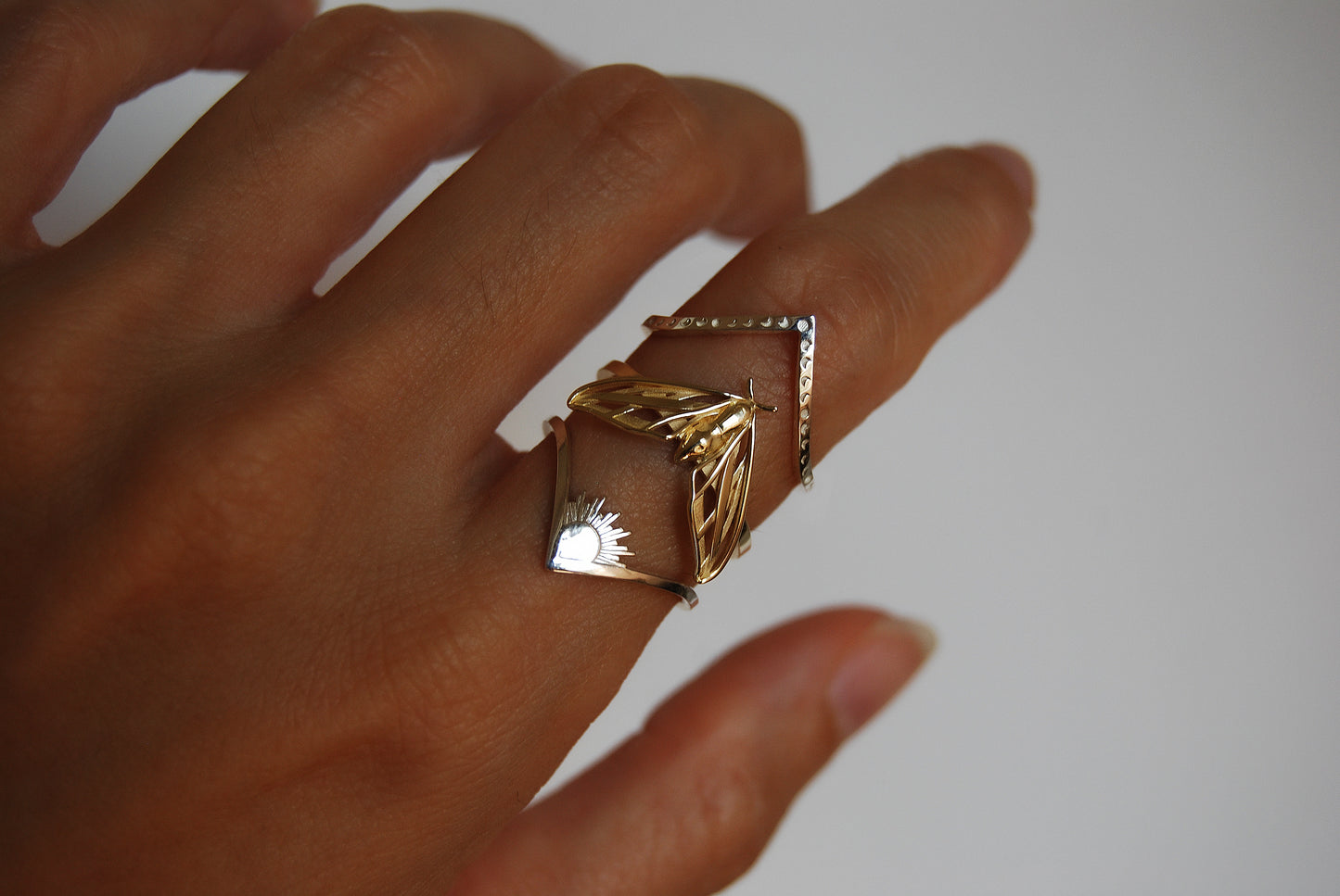 
Sterling silver sun and moon chevron rings stacked with gold moth ring on finger, blending mixed-metal celestial motifs.
