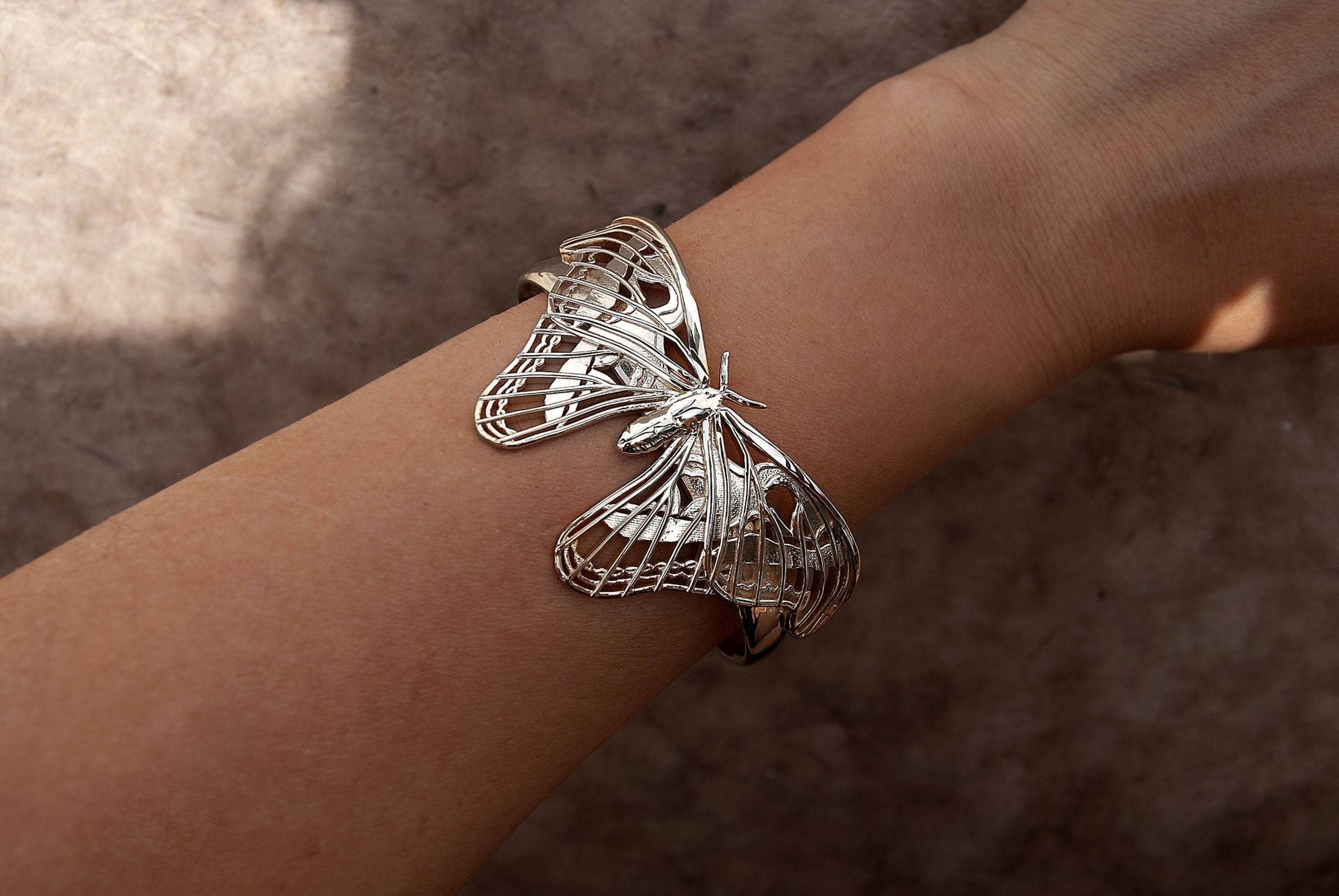 Close-up of the Atlas Moth Bracelet in sterling silver, worn on a hand. The detailed moth wings curve gracefully around the wrist, with a soft metallic sheen that highlights the sculptural design. The bracelet’s cutout elements and natural contours evoke elegance and craftsmanship inspired by nature.